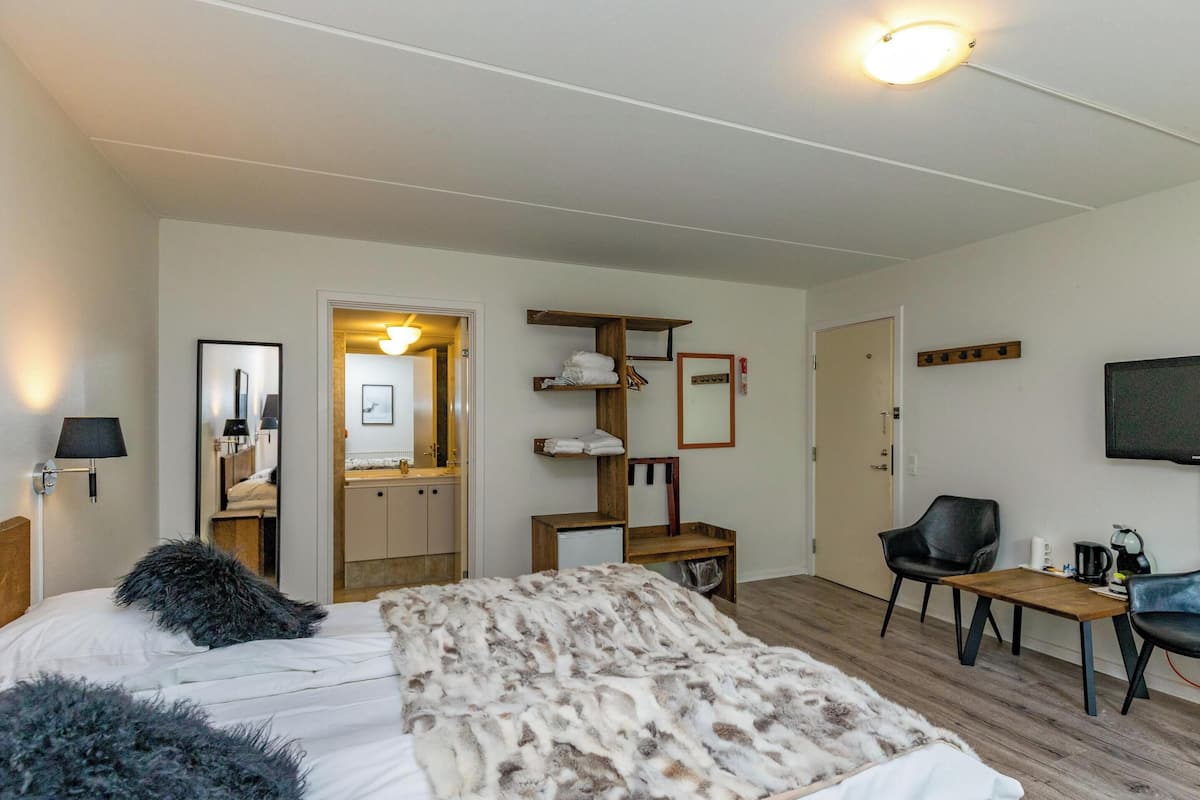 A bedroom with a bed, a wooden nightstand, a chair, a small table, a mirror, and a television.