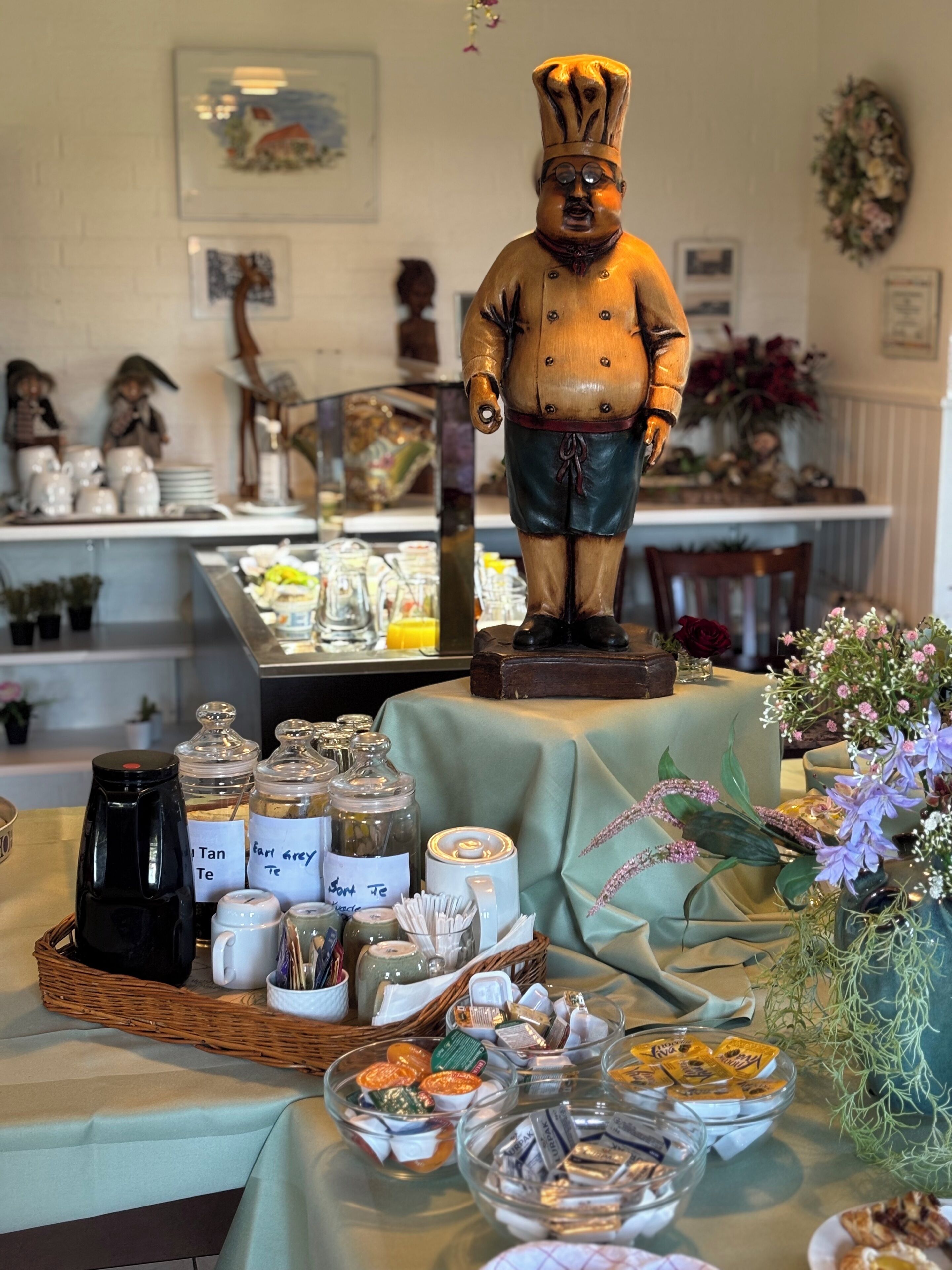 Una mesa de buffet con diversos alimentos y aderezos, una estatua decorativa de un chef y una disposición floral.