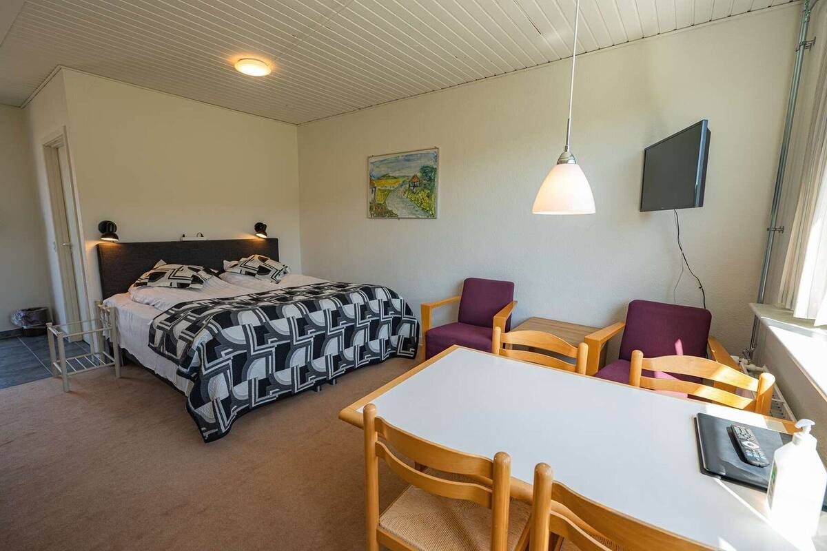 A bedroom with a bed, a desk with two chairs, a television, and a painting on the wall.