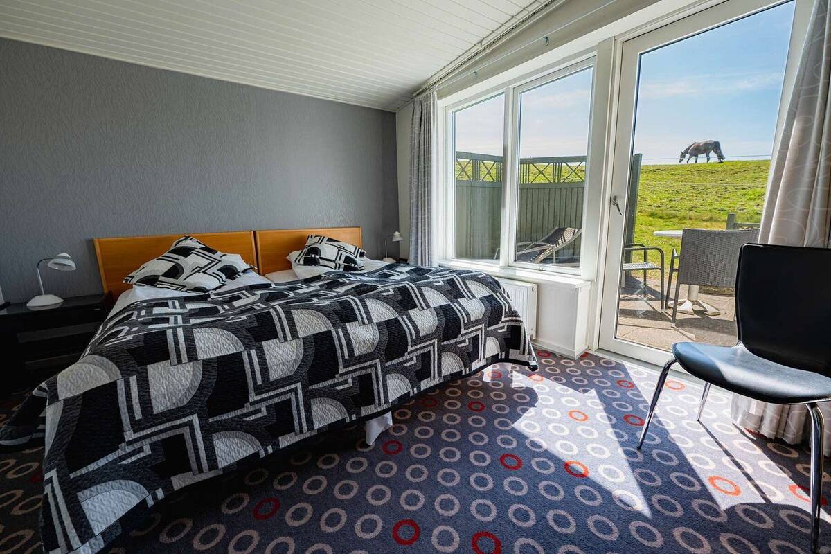 A bedroom with a bed, a nightstand, a chair, and a large window with a view of a grassy field and a horse.