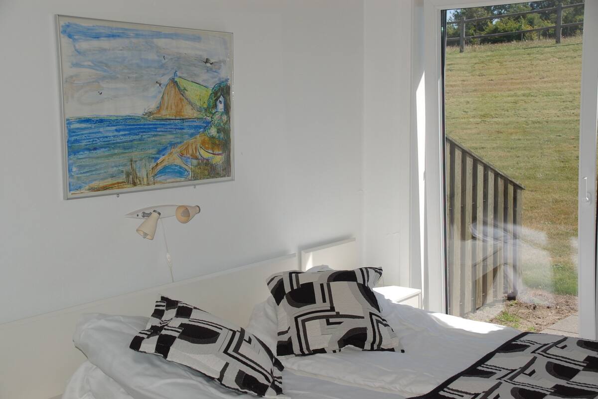 A bedroom with a bed, a painting, and a view of a grassy area through a window.