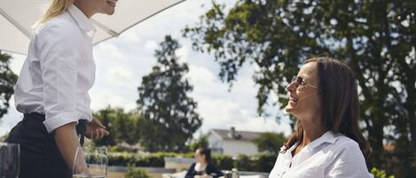 Un área de comedor al aire libre con una mesa preparada para dos personas, un camarero y un huésped.