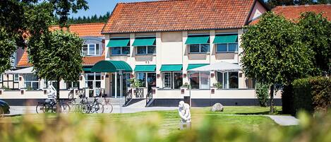 A building with a red-tiled roof, multiple windows, and a green awning. There are bicycles parked outside and a statue in the foreground.