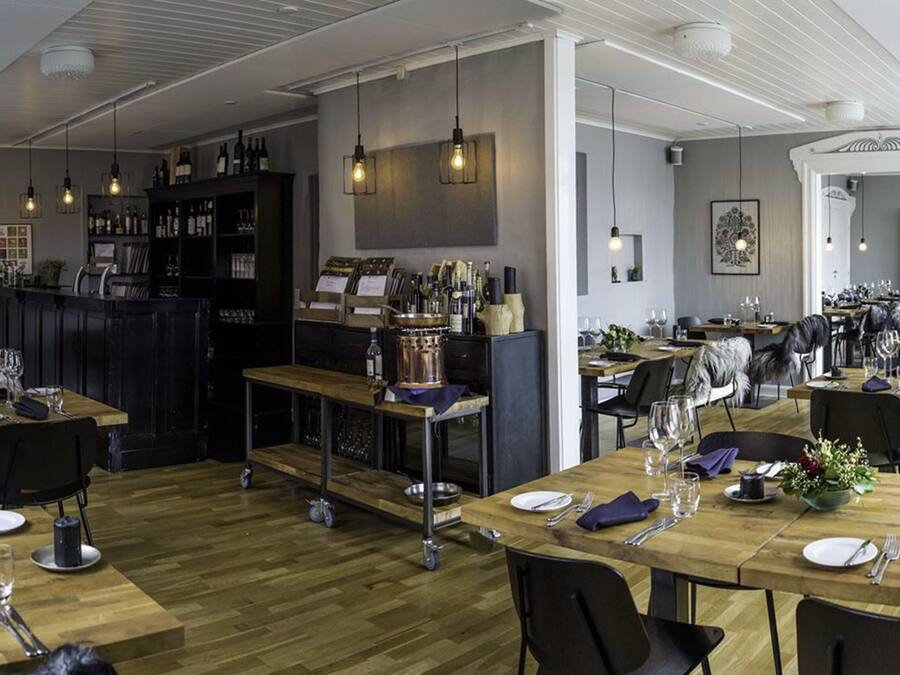 Un restaurante con mesas de madera, sillas negras y una zona de bar con botellas.