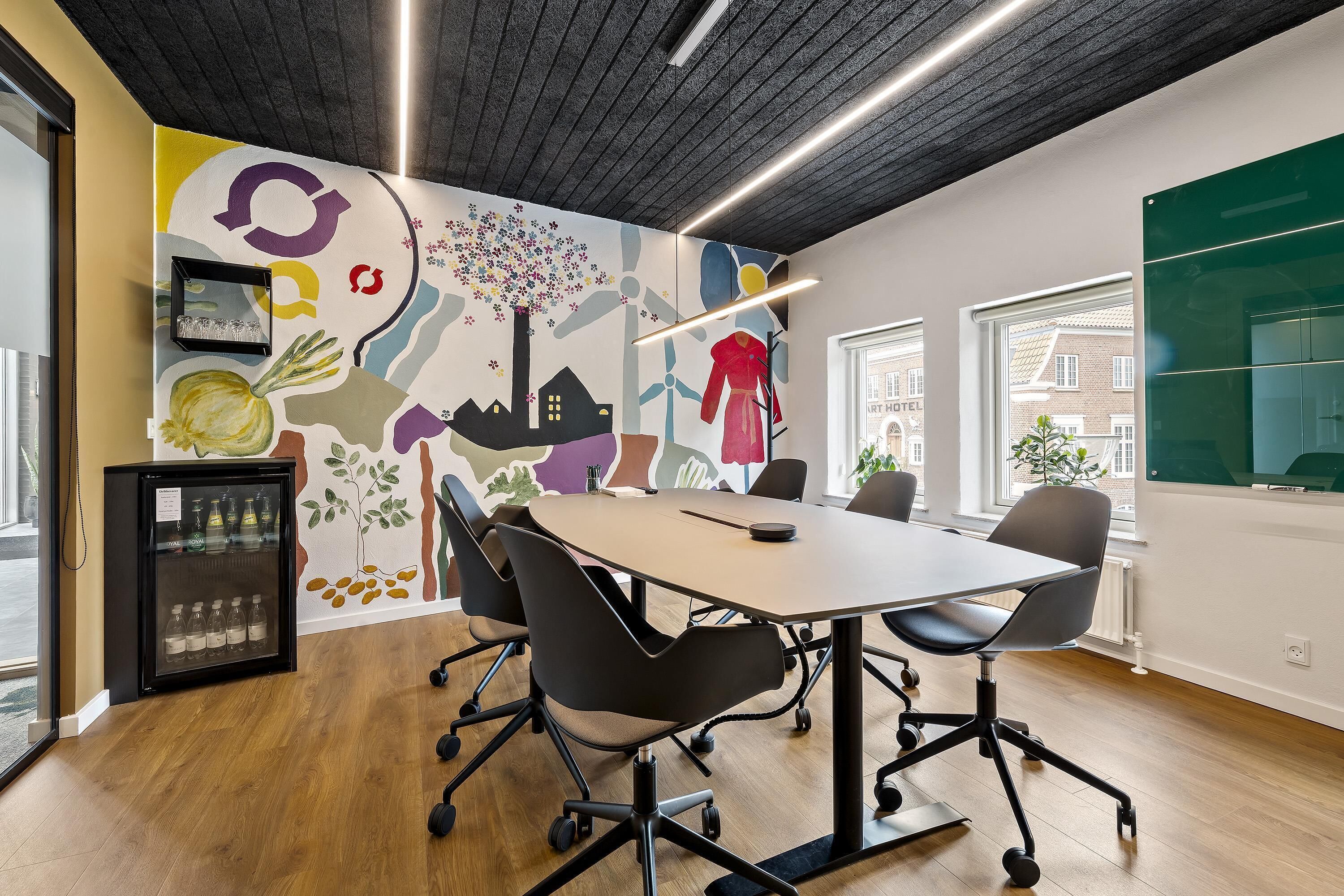 Un salón de conferencias con una gran mesa, sillas modernas, un mural en la pared y un refrigerador.