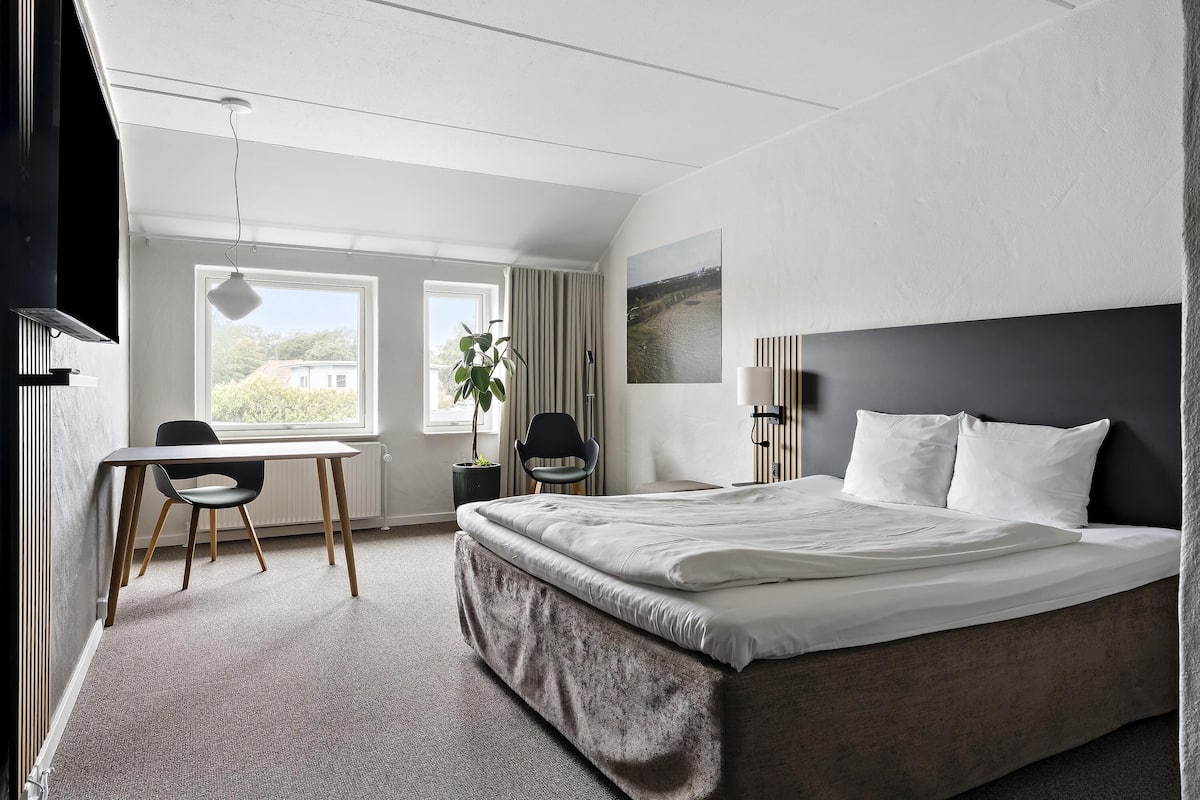 A hotel room with a bed, a desk, a chair, a plant, and a window with a view.