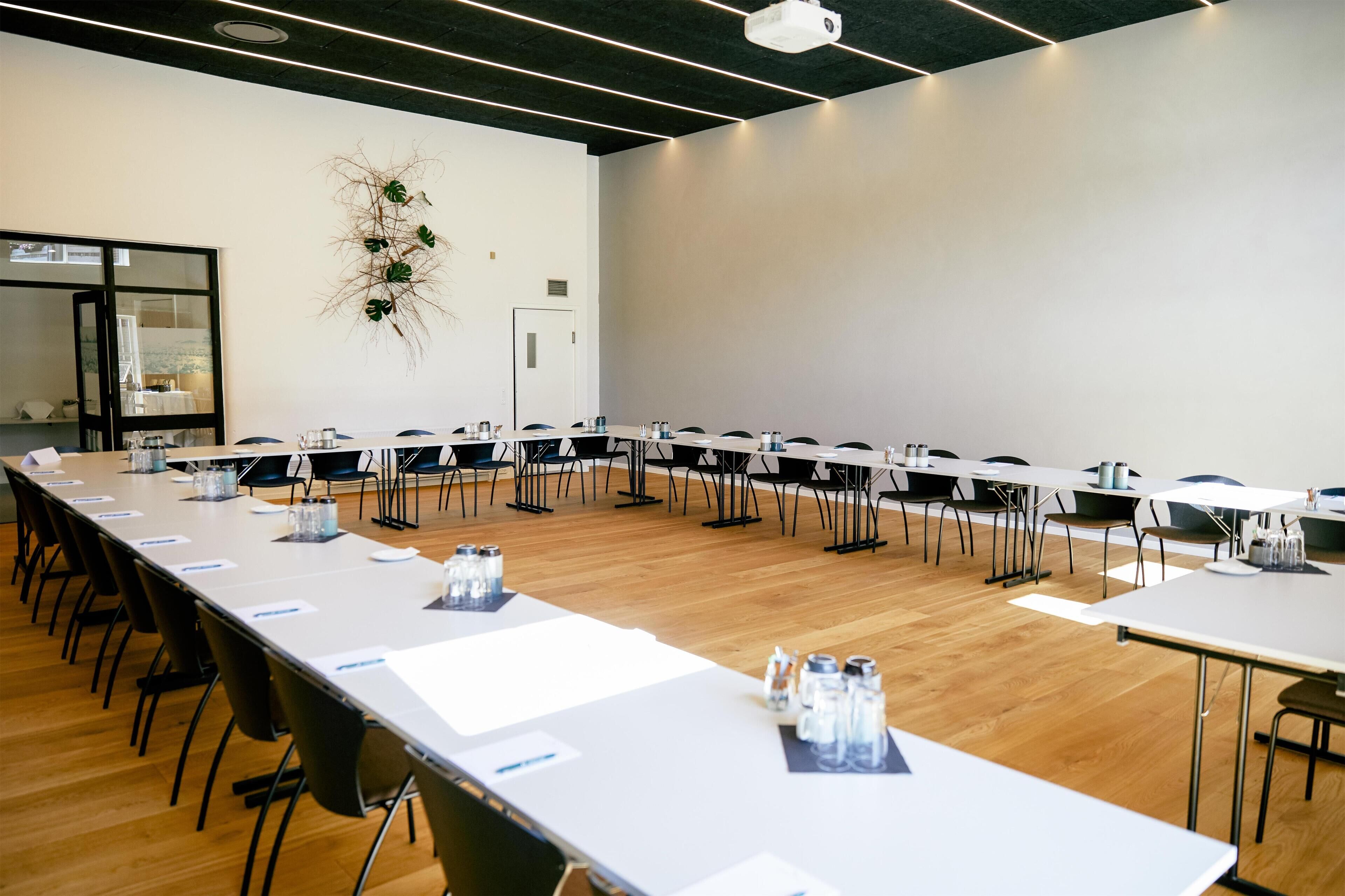 Un salón de conferencias con mesas largas y sillas, un proyector y una planta decorativa.