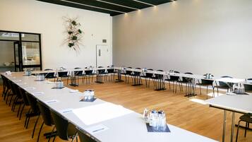 Un salón de conferencias con mesas largas y sillas, un proyector y una planta decorativa.
