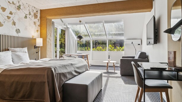 Une chambre d’hôtel moderne, dotée d’un grand lit, d’un bureau, d’une chaise et offrant une vue sur la verdure grâce à de grandes fenêtres.