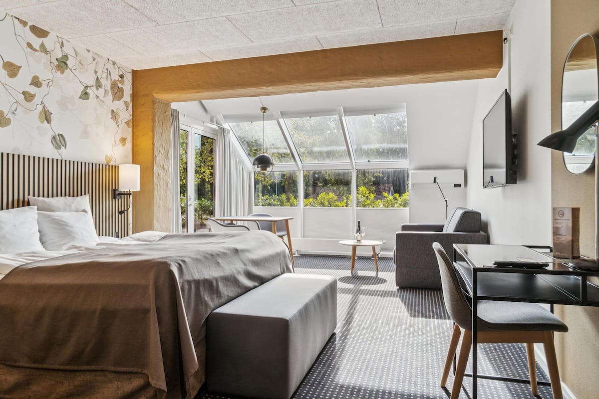 A modern hotel room with a large bed, a desk, a chair, and a view of greenery through large windows.