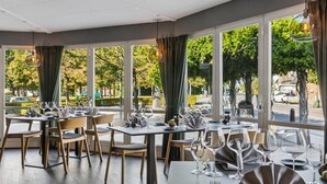 Un restaurant avec des tables dressées pour le service, de grandes fenêtres avec des rideaux et une vue sur la rue.