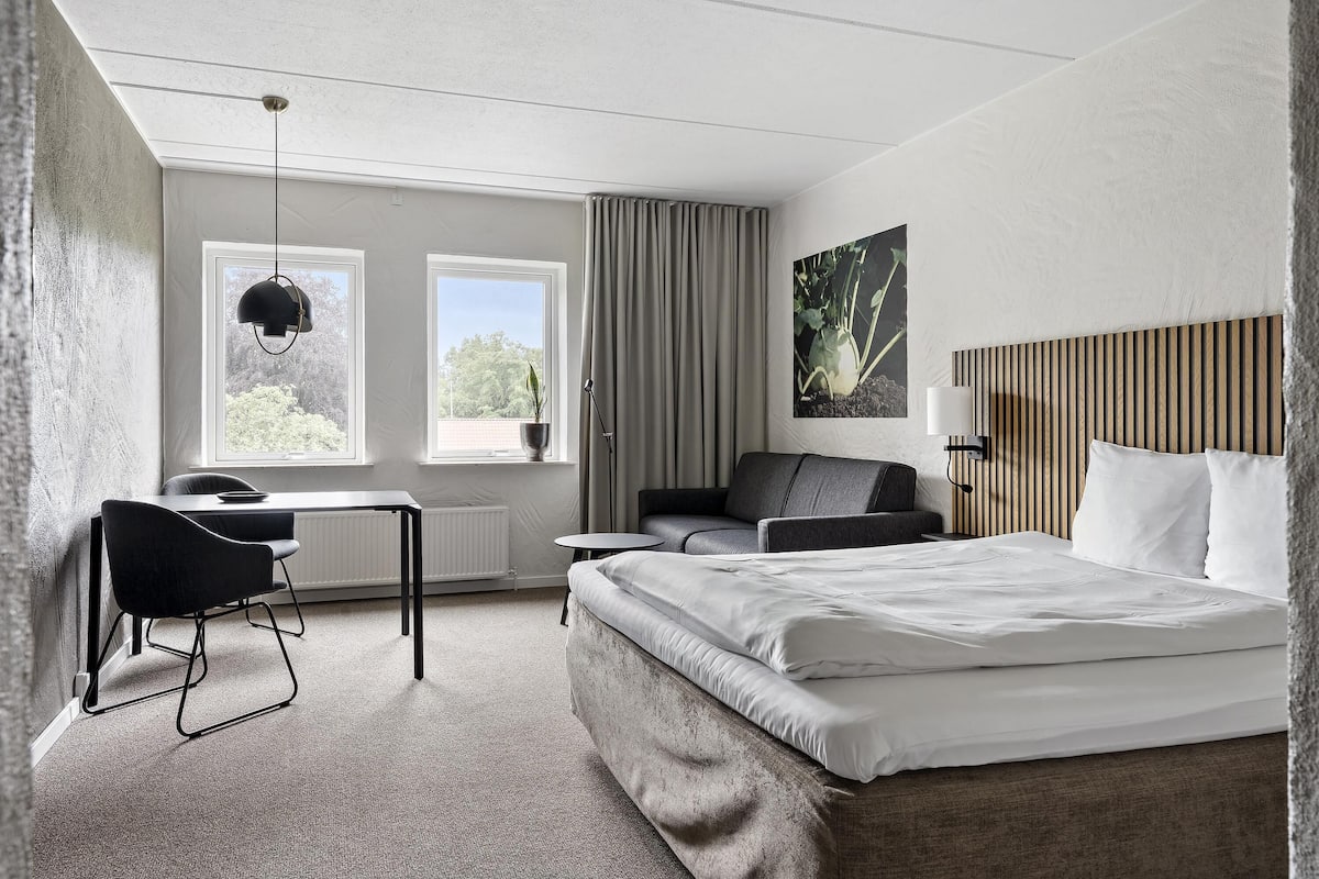 A hotel room with a bed, a desk, a chair, a small table, and a window with curtains.