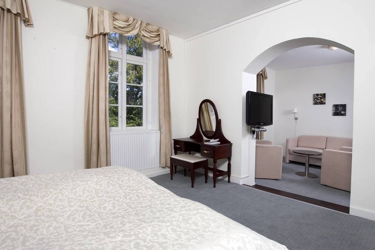 A hotel room with a large bed, a desk with a mirror, a television, a sofa, and a small table.