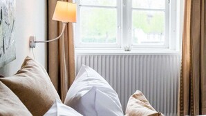 A bedroom with a bed, pillows, a lamp, and a window with curtains. - Gram Slotskro (Gram)