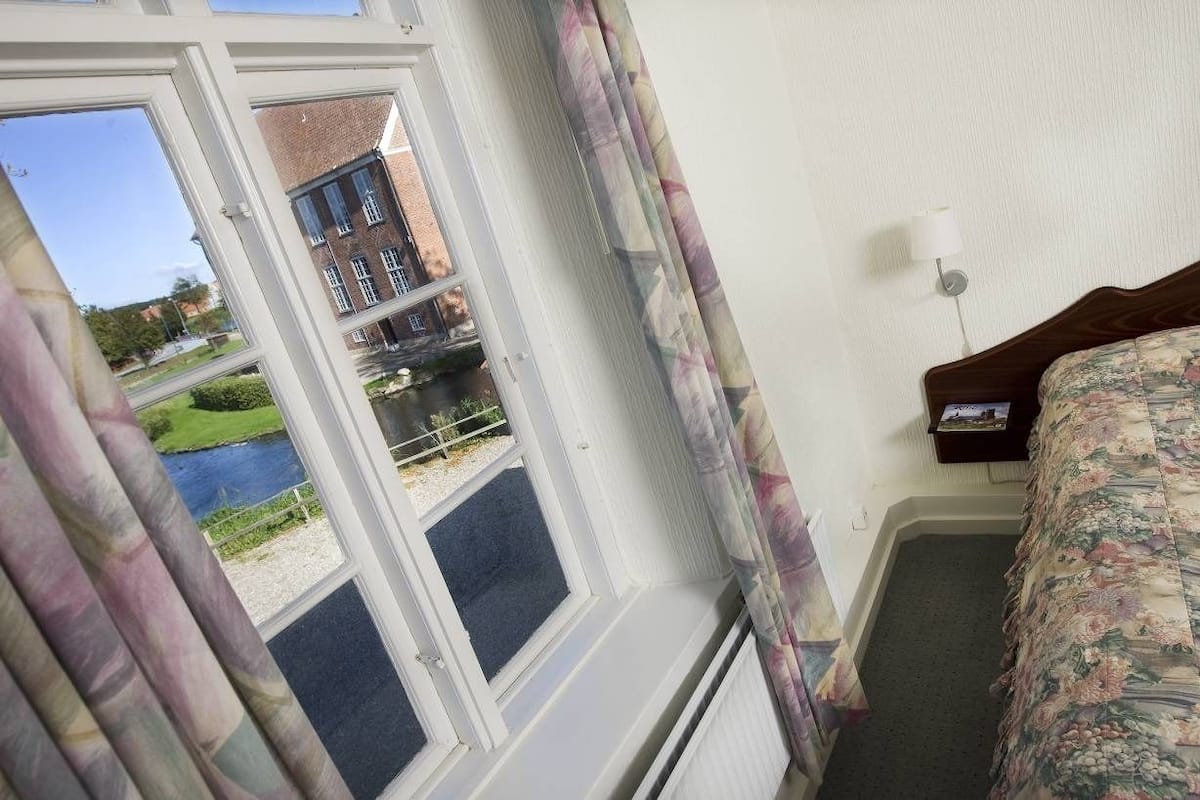 A hotel room with a bed, a nightstand, a window with curtains, and a view of a building and greenery outside.