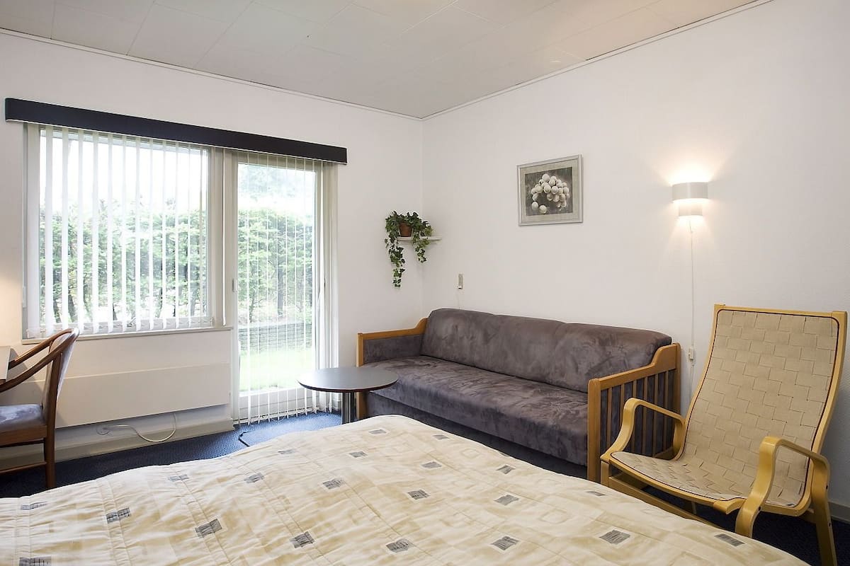 A hotel room with a bed, a sofa, a chair, a small table, a window with blinds, a wall-mounted light, and a framed picture.