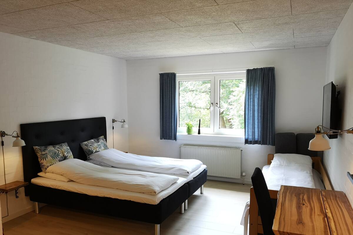 Quarto de hotel com uma cama grande, uma cama menor, uma mesa de madeira e uma janela com cortinas.
