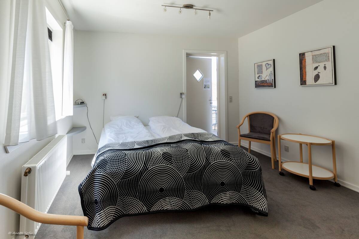 Quarto com uma cama, uma cadeira, uma mesa pequena e duas obras de arte na parede.