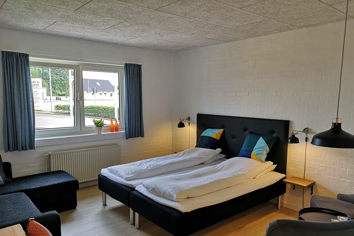 Quarto de hotel com uma cama grande, uma escrivaninha, uma cadeira e uma janela com cortinas.