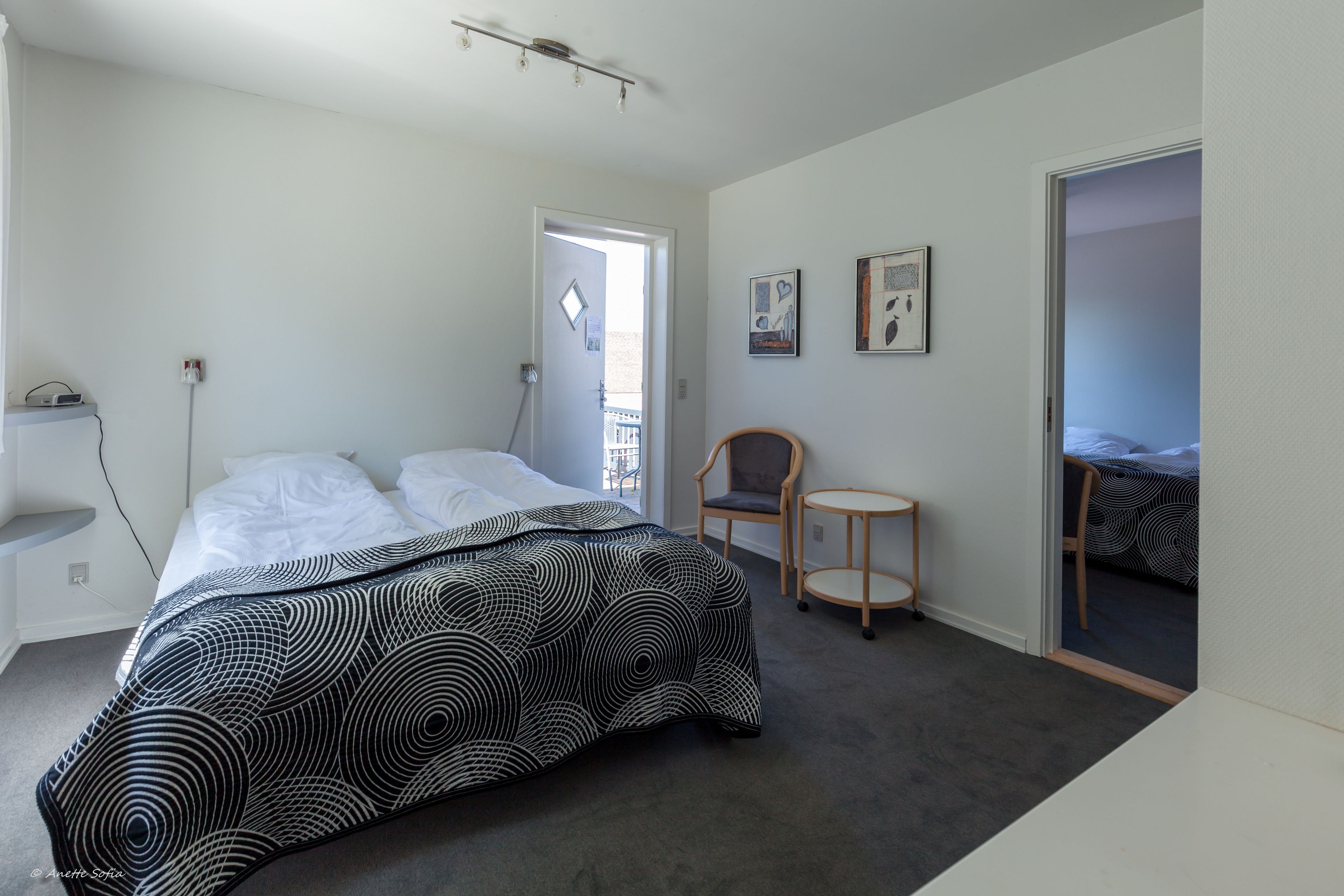 Un dormitorio con cama, una silla, una mesita y una puerta que da a otra habitación.