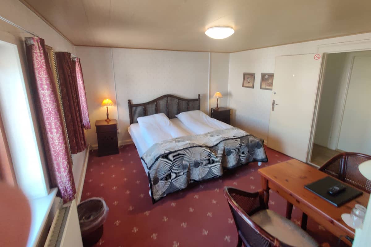 Quarto de hotel com uma cama, duas mesas de cabeceira com abajures, uma escrivaninha de madeira, uma cadeira e uma porta.