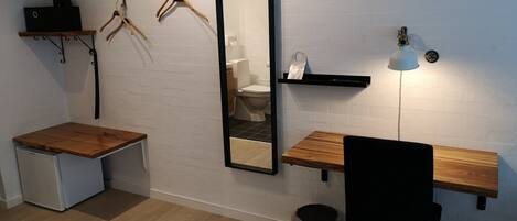 Habitación de hotel pequeña con escritorio de madera, espejo, lavamanos y refrigerador compacto.