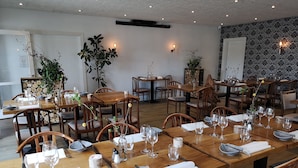 Un restaurante con mesas y sillas de madera, dispuesto en un amplio comedor con plantas en macetas e iluminación en la pared.