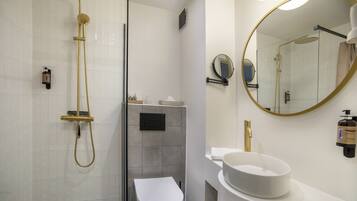 Un baño moderno con inodoro blanco, ducha con cabezal dorado, espejo redondo y lavamanos blanco.