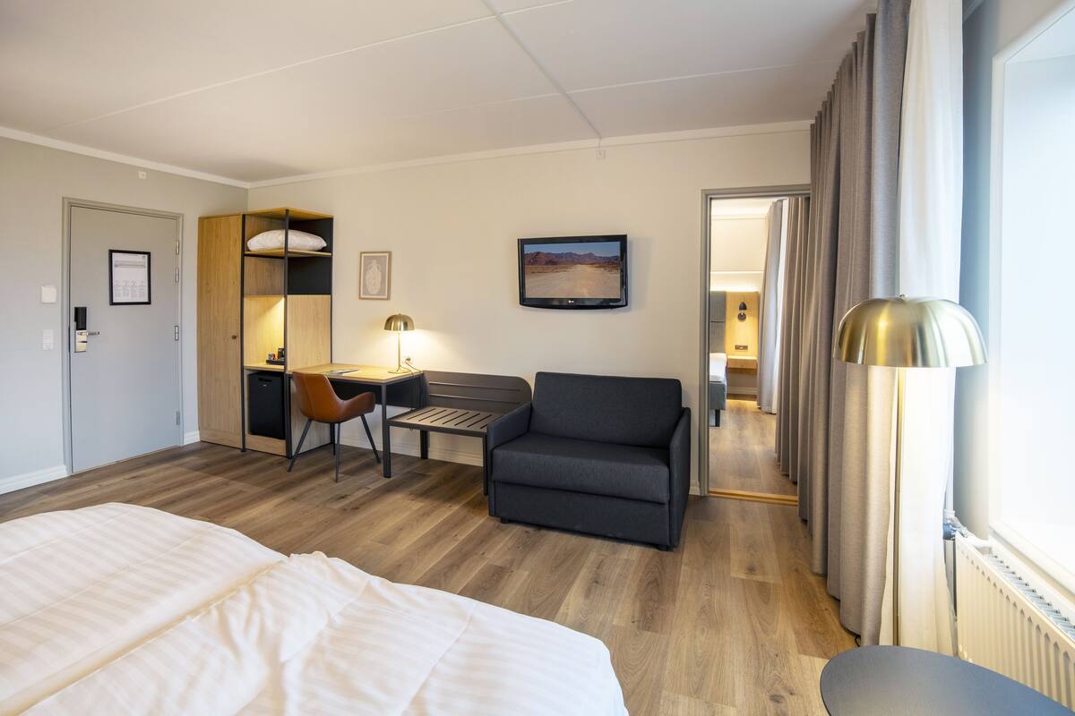 Habitación de hotel con cama, escritorio, silla, sofá, televisor y ventana con cortinas.