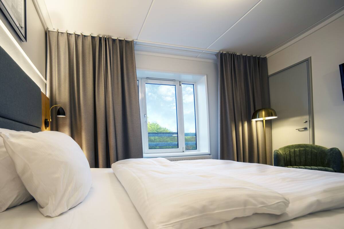 Una habitación de hotel con cama, ventana con cortinas, lámpara y una silla.