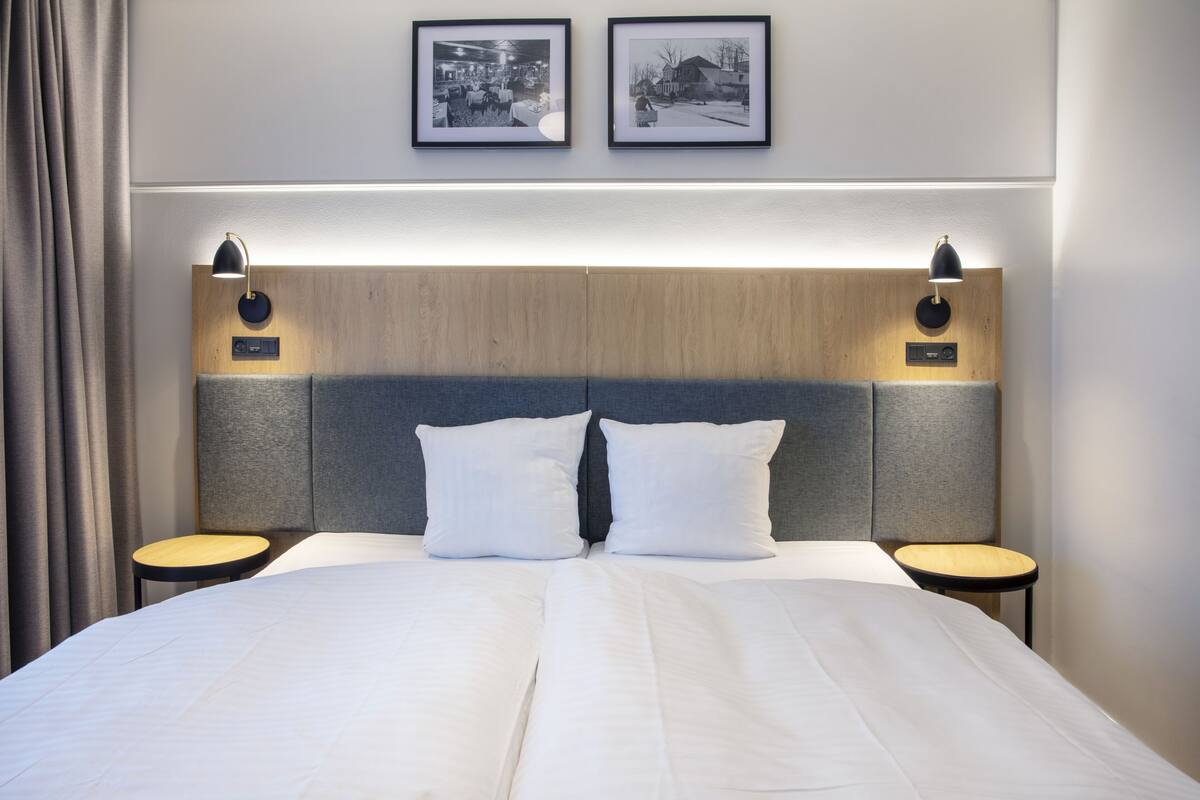 Una cama bien hecha con sábanas blancas, dos mesitas de noche con lámparas, y dos cuadros enmarcados en la pared de arriba.