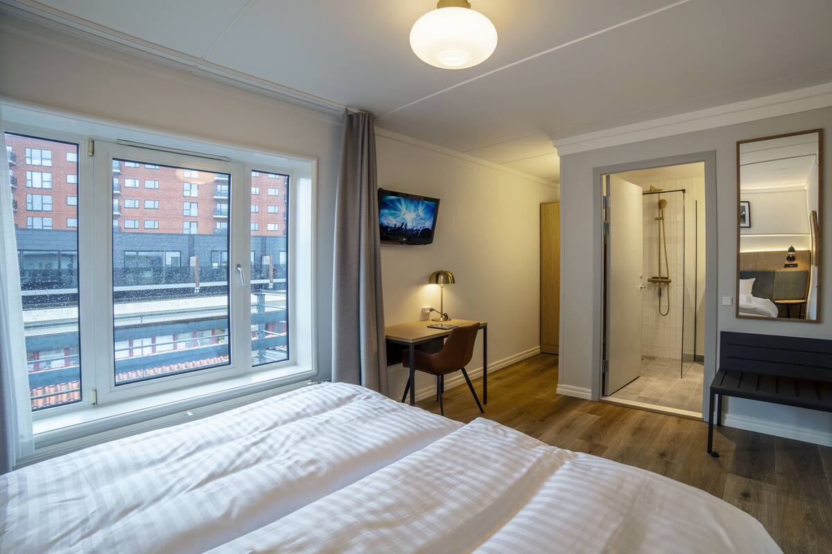 Habitación de hotel con una cama grande, un escritorio con silla, televisión, zona de ducha y vistas a un edificio por la ventana.