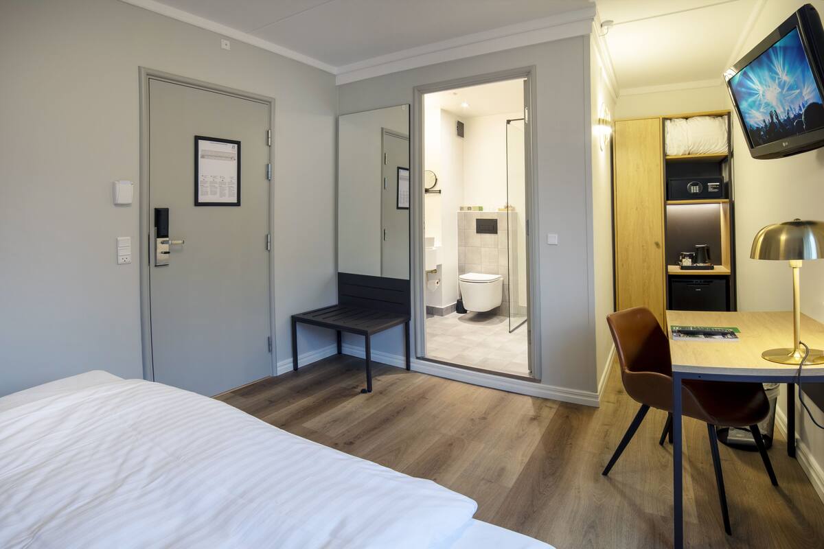 Habitación de hotel con cama, escritorio y silla, televisor y baño visible a través de una puerta abierta.