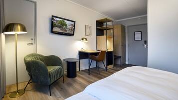 A hotel room with a bed, a desk with a chair, a green armchair, a floor lamp, and a wall-mounted TV.