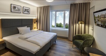 A modern hotel room with a large bed, a green armchair, a bedside table, a lamp, and a window with curtains.