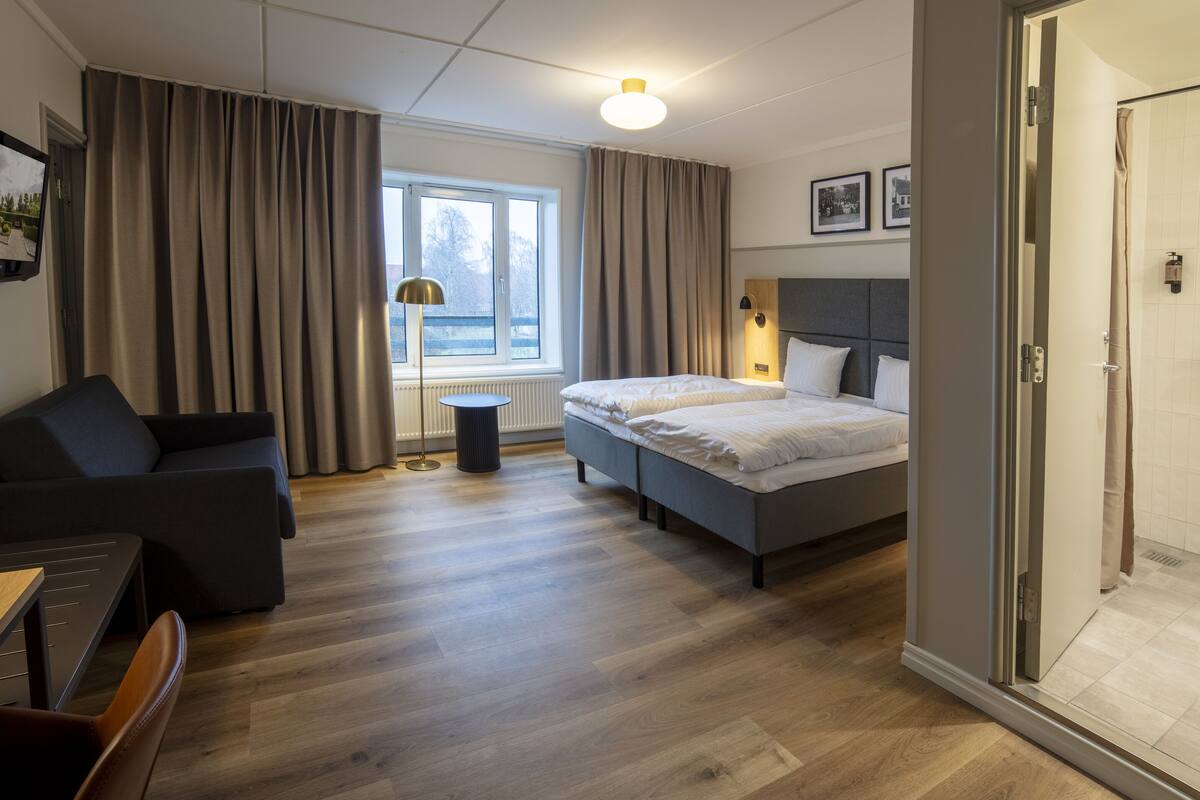 Una habitación de hotel moderna con cama, escritorio, silla, sofá, ventana con cortinas, lámpara de noche y zona de ducha.