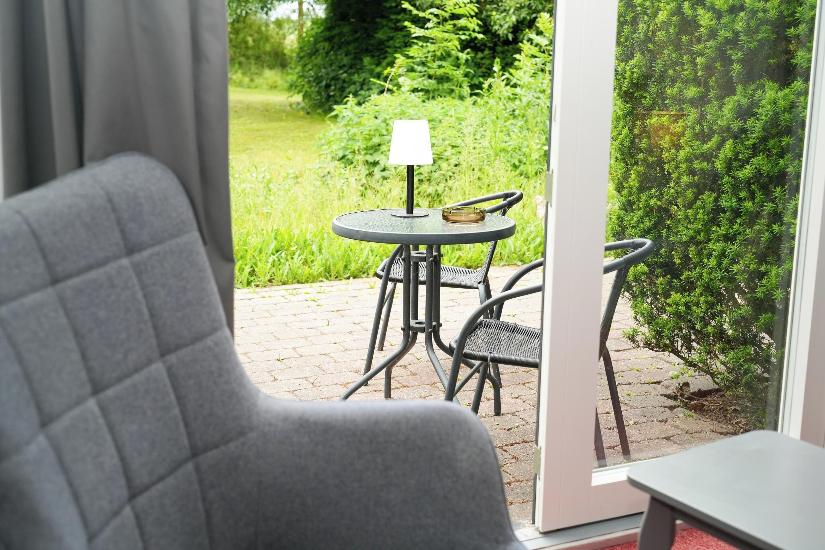 En vy från ett rum med en grå klädd fåtölj, ett litet runt bord med en lampa och en glasdörr som leder ut till en uteplats med ett bord och stolar.