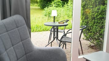 En vy från ett rum med en grå klädd fåtölj, ett litet runt bord med en lampa och en glasdörr som leder ut till en uteplats med ett bord och stolar.
