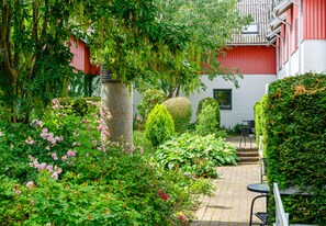 Experience a serene garden setting with lush greenery and blooming flowers, surrounded by a well-maintained structure and inviting seating areas.