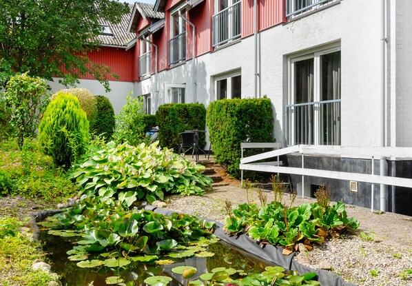 Un patio con un estanque, abundante vegetación y un edificio con paredes exteriores rojas y blancas.