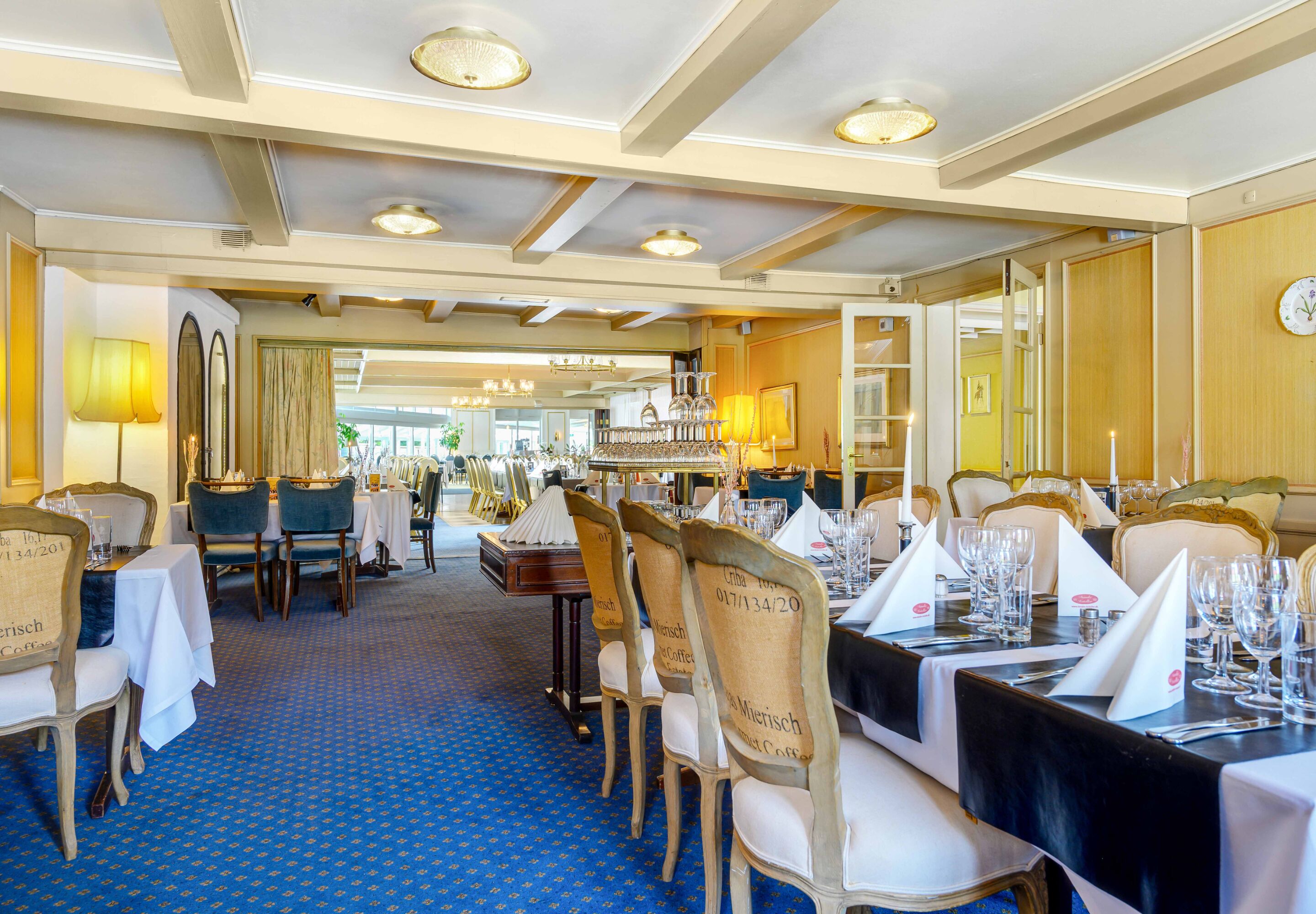 A formal dining room with tables set for a meal, chairs with labels, and a blue carpeted floor.