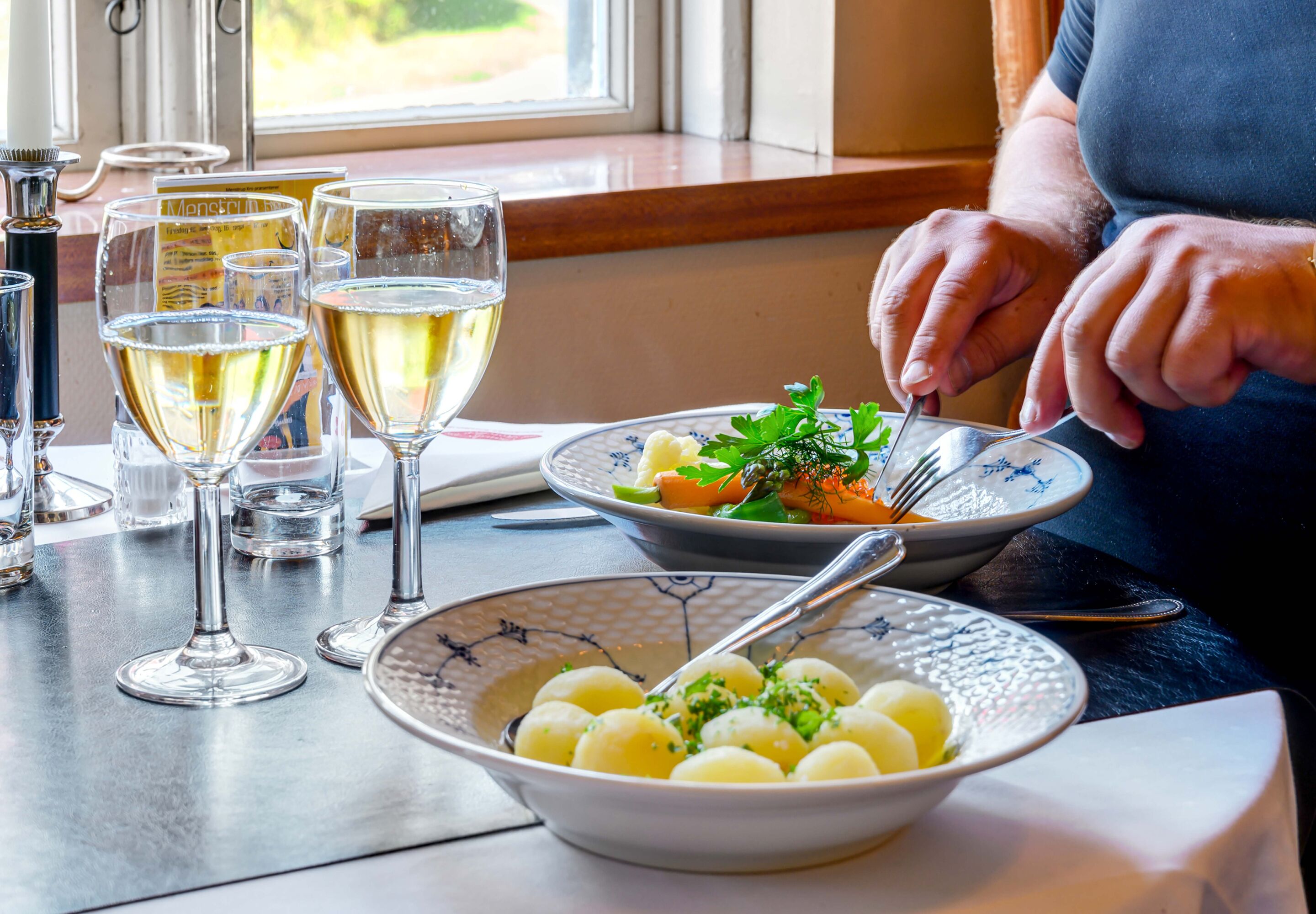 A dining table with a plate of food, a bowl of potatoes, and several glasses of white wine.