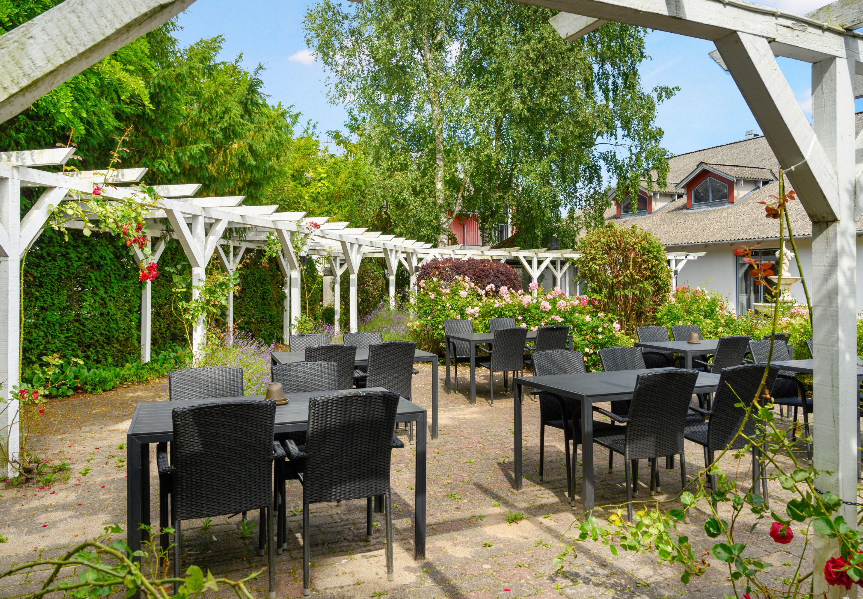 An outdoor patio with tables and chairs, surrounded by lush greenery and a white pergola structure.