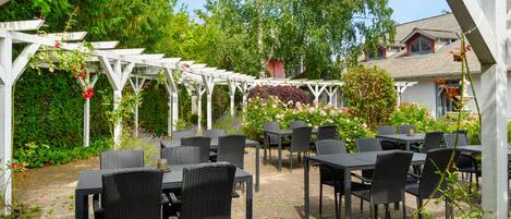 An outdoor patio with tables and chairs, surrounded by lush greenery and a white pergola structure.