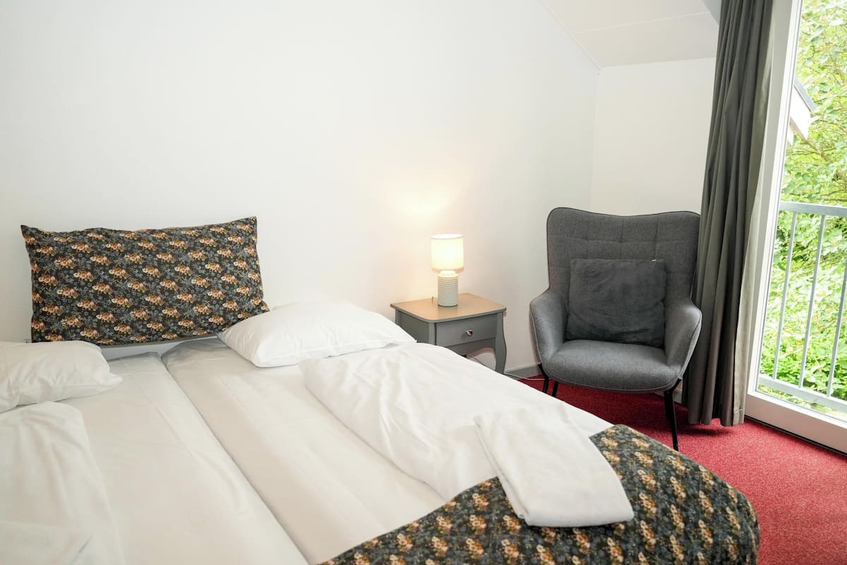 A hotel room with a bed, a chair, a nightstand with a lamp, and a window with curtains.