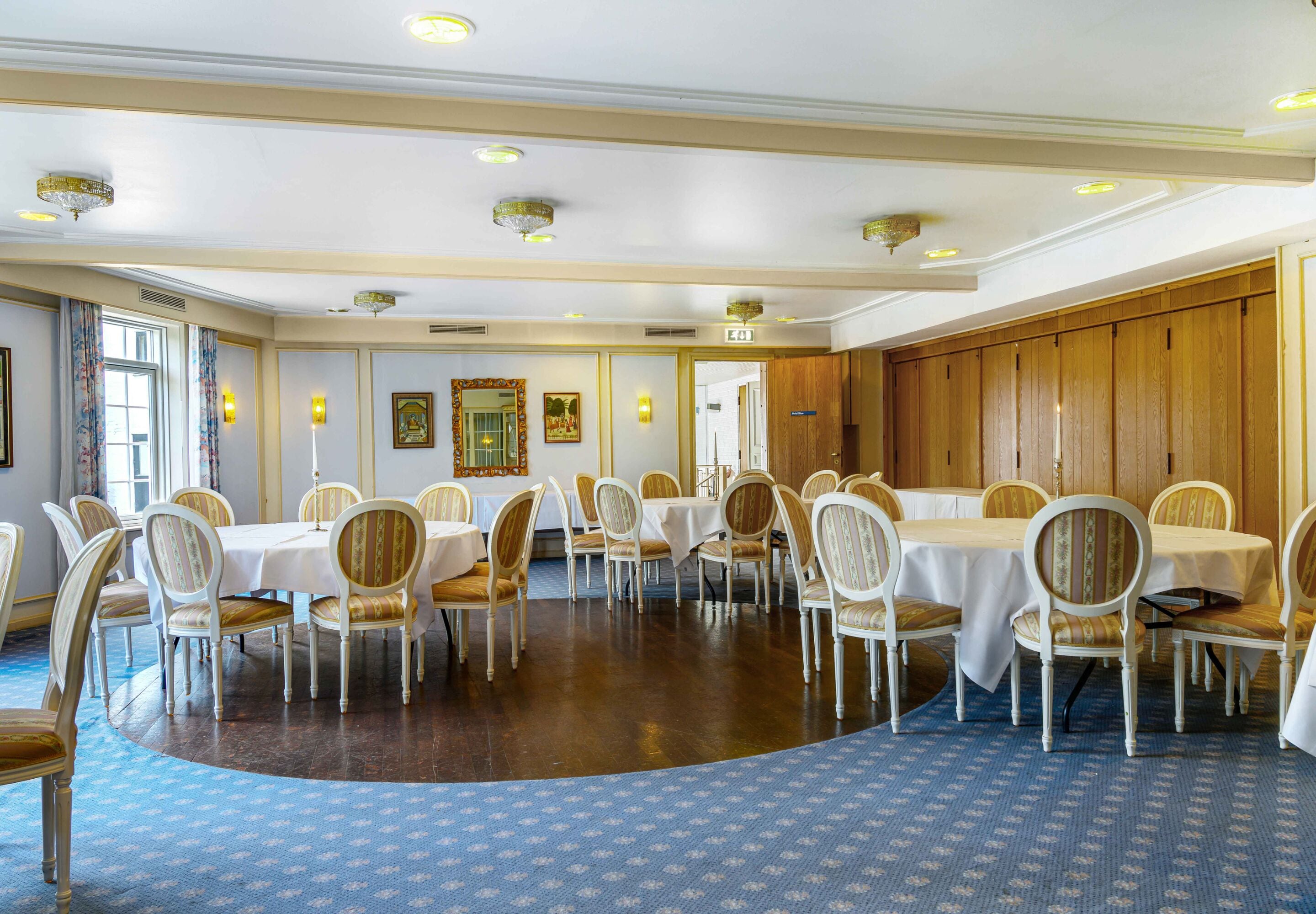 A large, well-lit dining room with round tables and chairs, wooden paneling on the walls, and framed artwork on the walls.