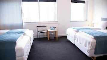 A hotel room with two beds, a small table, a chair, and two windows.