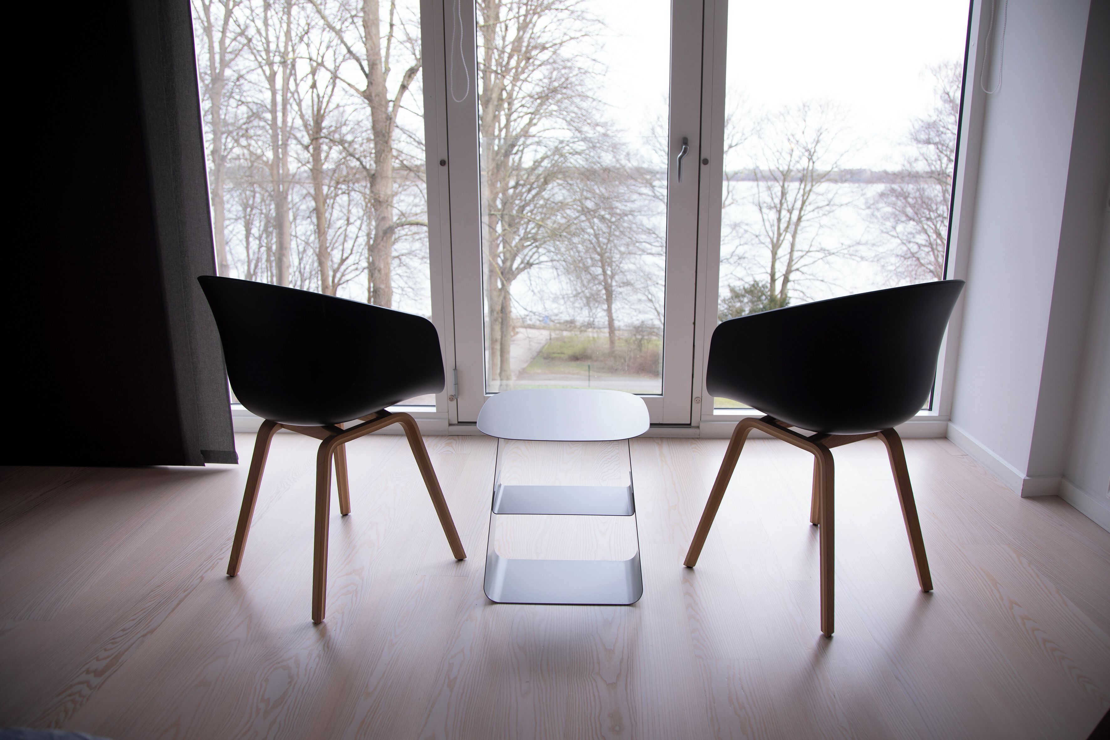 A modern room with two black chairs and a small white table, large windows with a view of trees and a body of water.