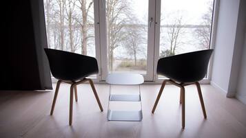 Una habitación moderna con dos sillas negras y una mesita blanca, ventanas amplias con vistas a árboles y un cuerpo de agua.