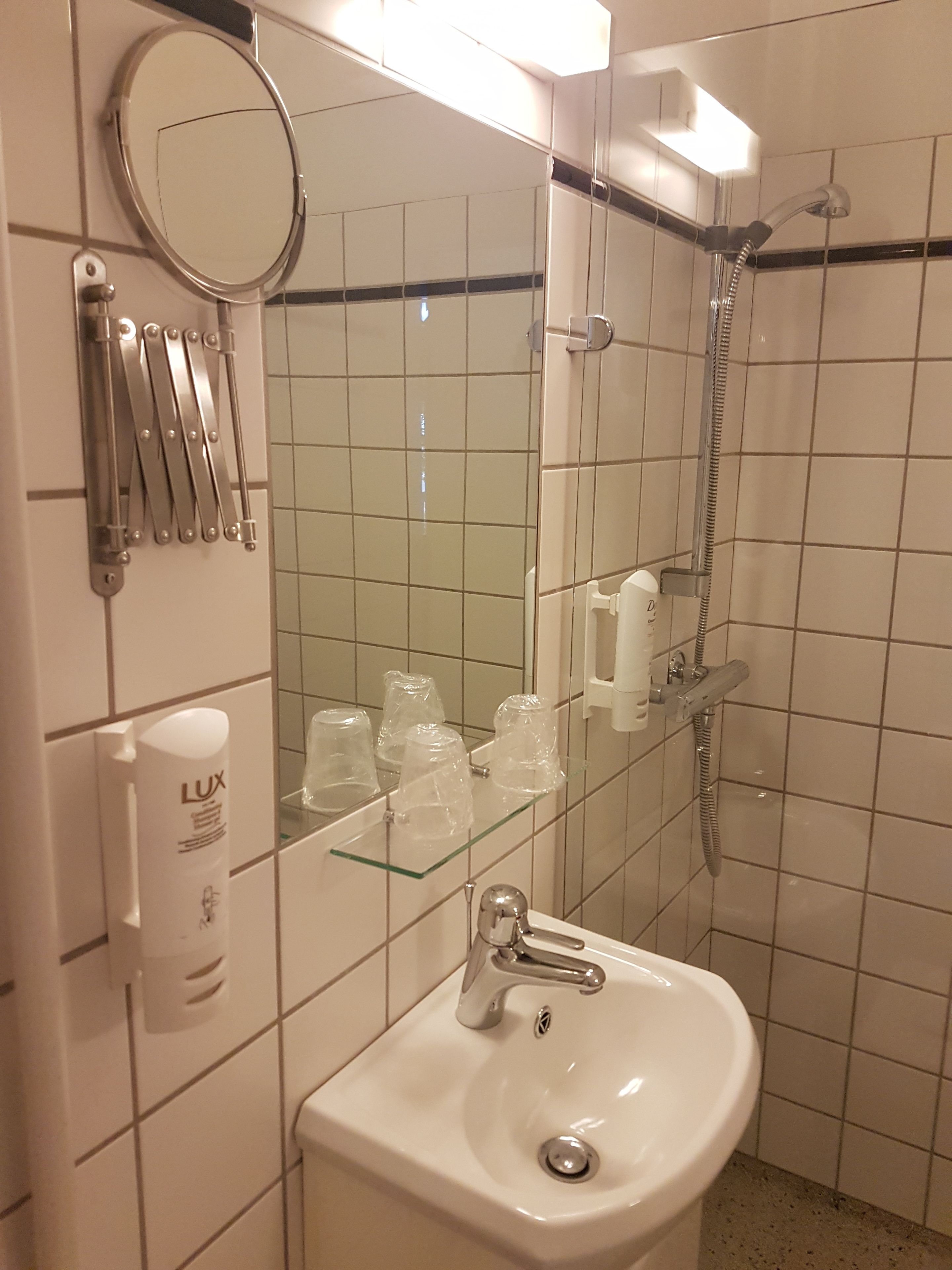 A bathroom with a sink, mirror, and shower. There are toiletries on the wall and a glass shelf.